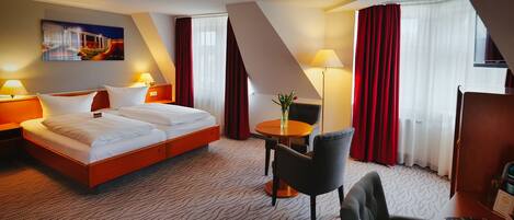 Een hotelkamer met een groot bed, een bureau, een stoel, een kleine tafel met een vaas, en een schilderij aan de muur.
