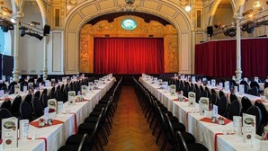Un salón de banquetes con techo ornamentado y candelabros, filas de mesas con tarjetas de nombres y un escenario con cortina roja.