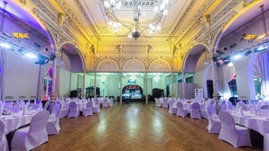 Un salón imponente con techo ornamentado y candelabros, mesas dispuestas con manteles blancos y sillas, y un escenario al frente.
