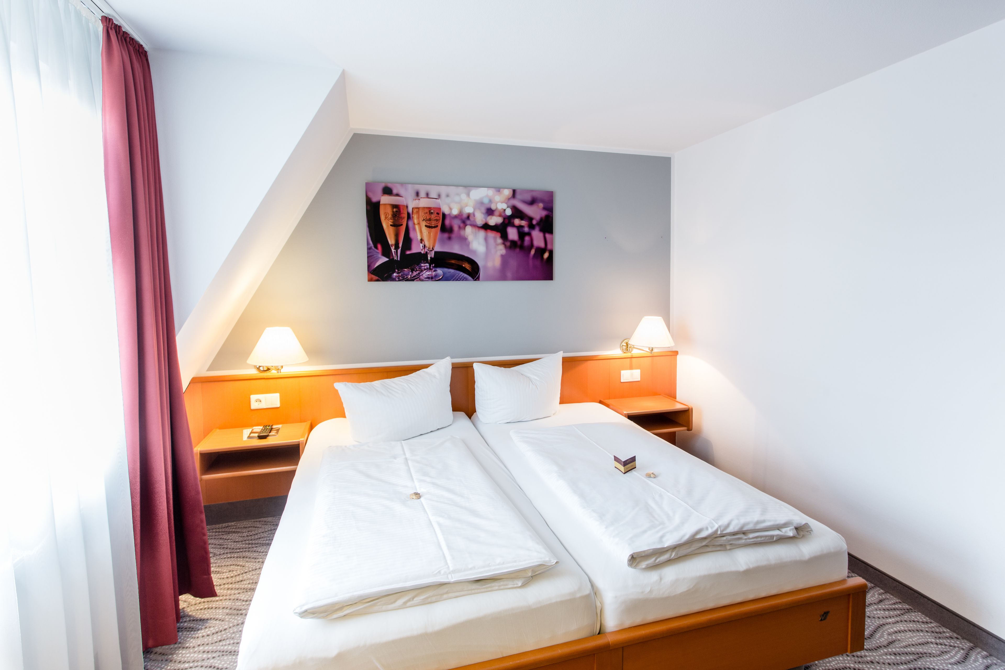 A hotel room with a double bed, two bedside tables, a nightstand, a wall-mounted picture, and a window with curtains.