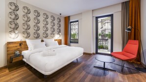 A modern hotel room with a large bed, a red chair, a small table, and a balcony with a view of buildings. - Shota@Rustaveli Boutique hotel (Tbilisi)