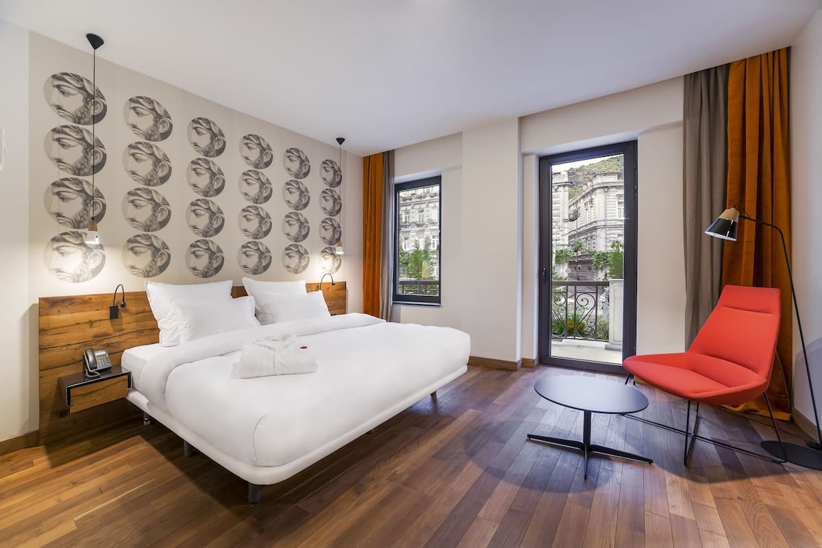 Ein modernes Hotelzimmer mit einem großen Bett, einem roten Sessel, einem kleinen Tisch und einem Balkon mit Blick auf Gebäude.