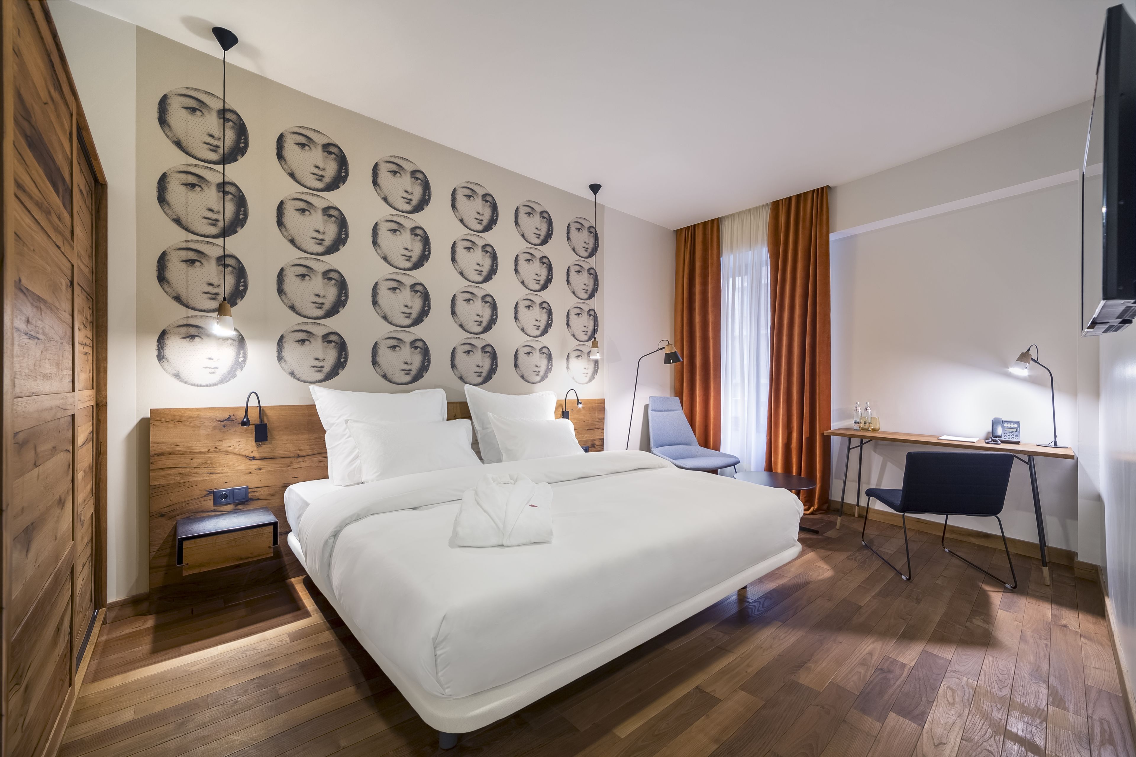 A hotel room with a large bed, a desk, a chair, and a wall with multiple face patterns.