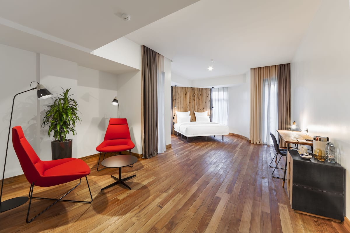 Ein Hotelzimmer mit Holzboden, einem Bett, einem Schreibtisch, zwei roten Stühlen, einer Topfpflanze und einem schwarzen Nachttisch.