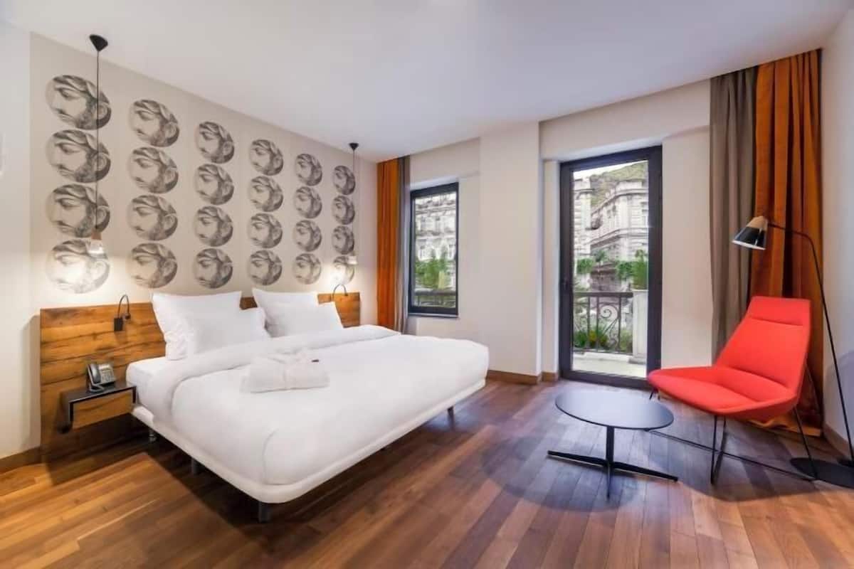 Ein Hotelzimmer mit Bett, einem roten Sessel, einem kleinen Tisch und einem Balkon mit Blick.