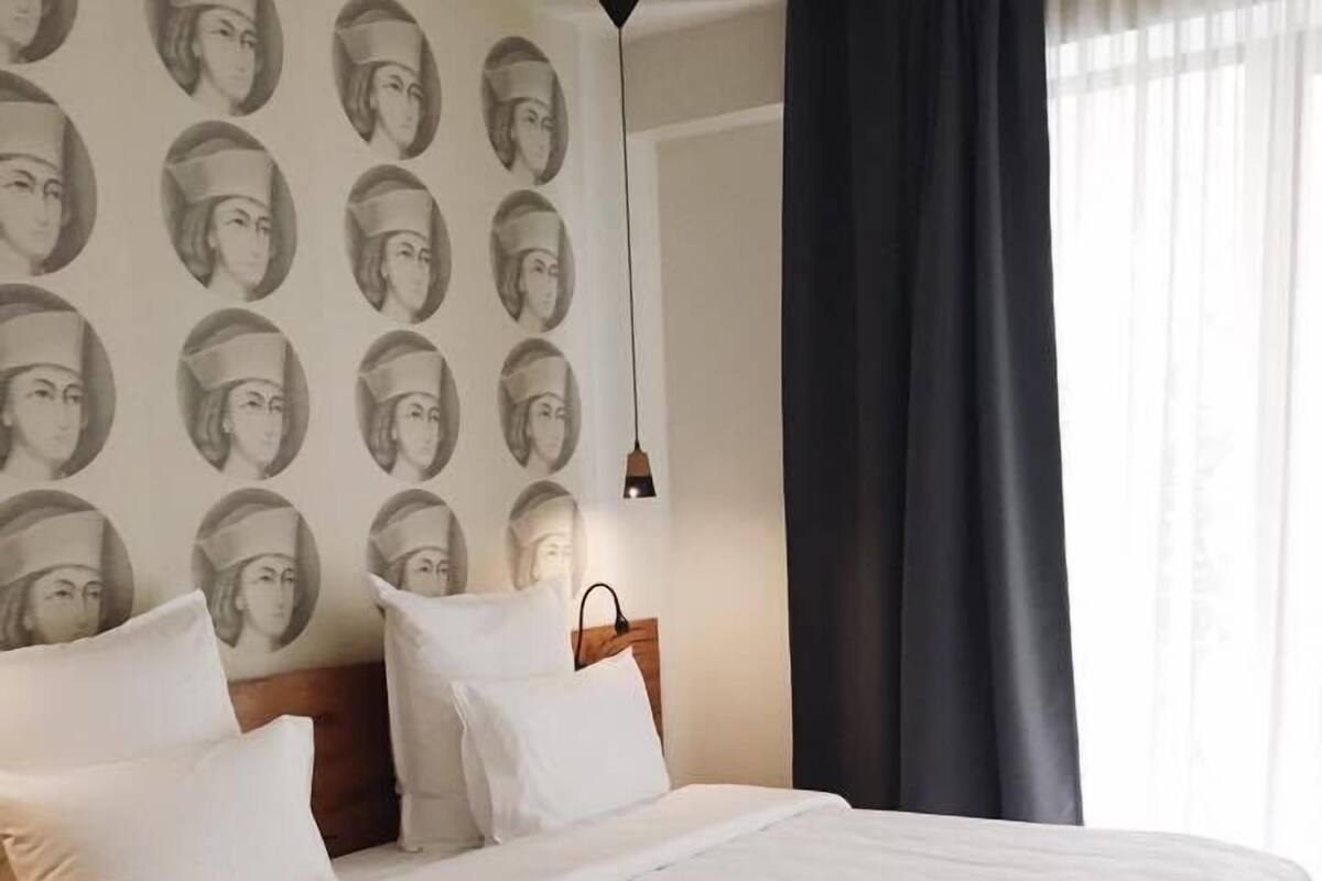 Ein Hotelzimmer mit einem großen Bett, einem Kopfteil, einem Nachttisch und einer Wand mit einem sich wiederholenden Muster aus Porträts.