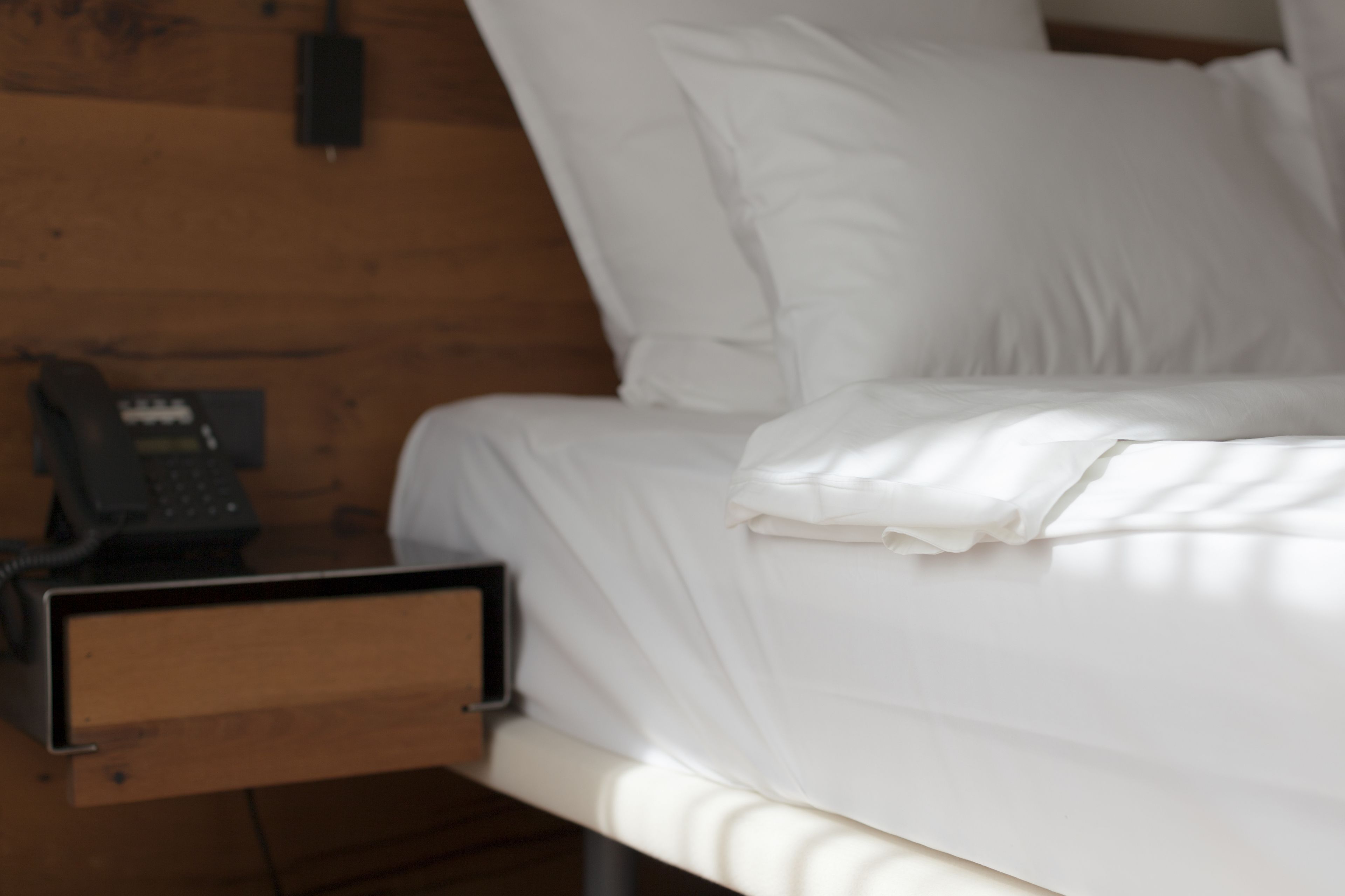 Una cama con sábanas blancas, una mesita de noche de madera con un teléfono y un cajón.
