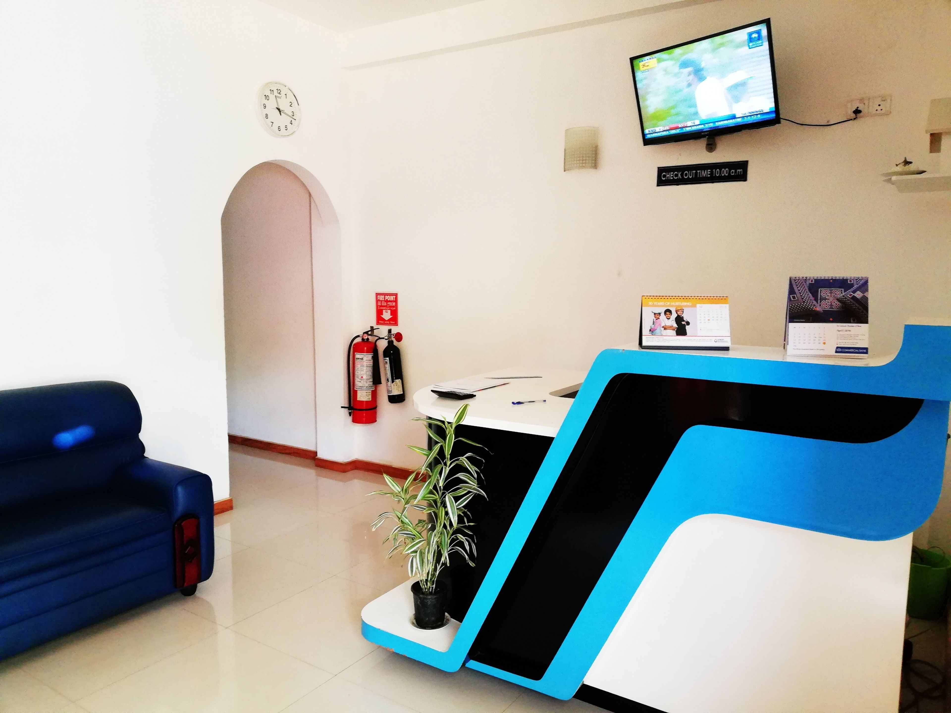 A reception area with a blue sofa, a reception desk, a fire extinguisher, a clock, and a television mounted on the wall.