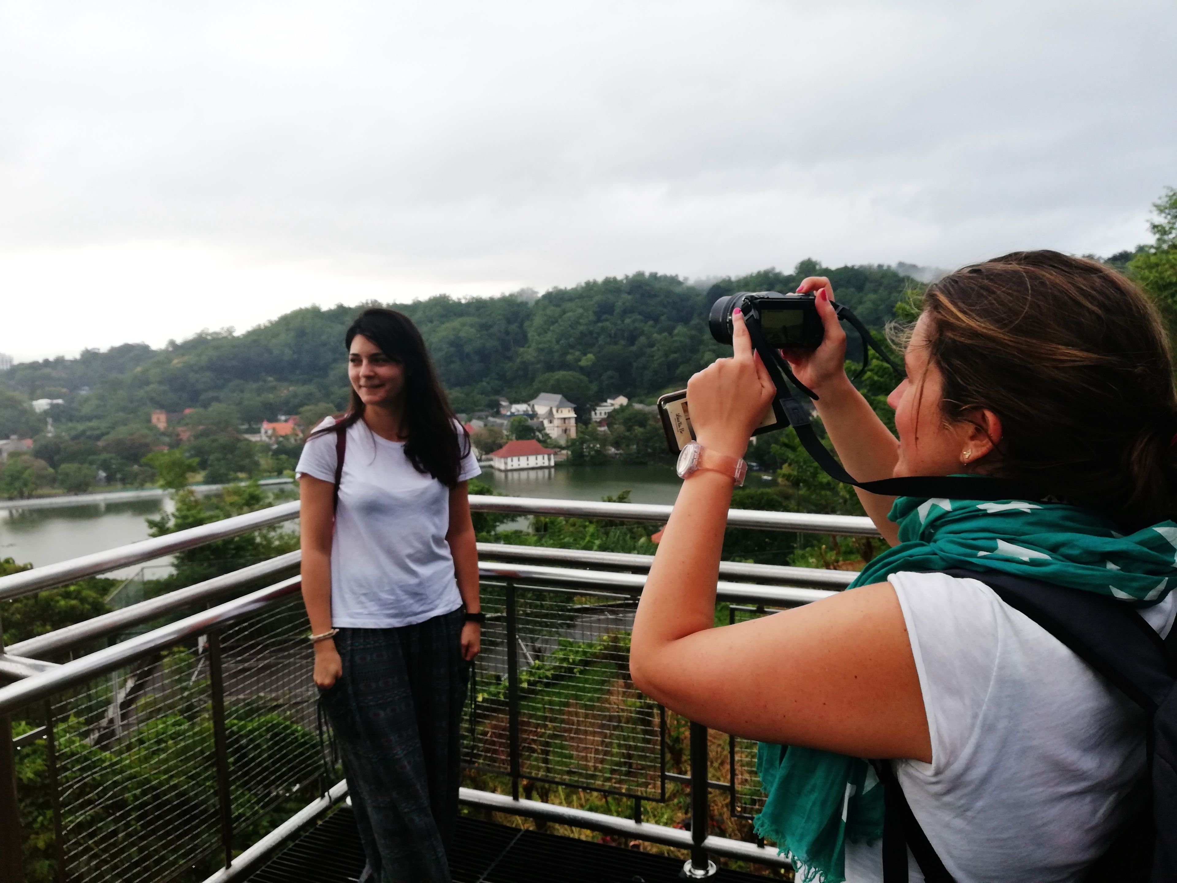 Una persona fotografa un'altra persona in piedi su un ponte, con un fiume e degli edifici sullo sfondo.