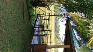 Un área de estar al aire libre techada, con estructura de madera y rodeada de césped y palmeras.