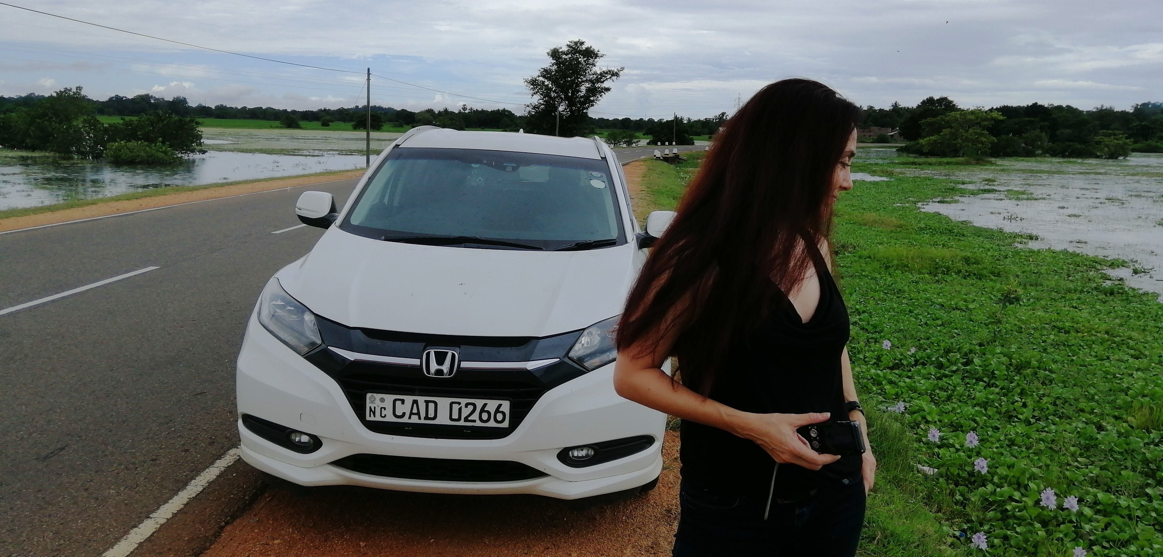 Une voiture Honda blanche est garée sur le bord d’une route, et une femme se tient à côté.