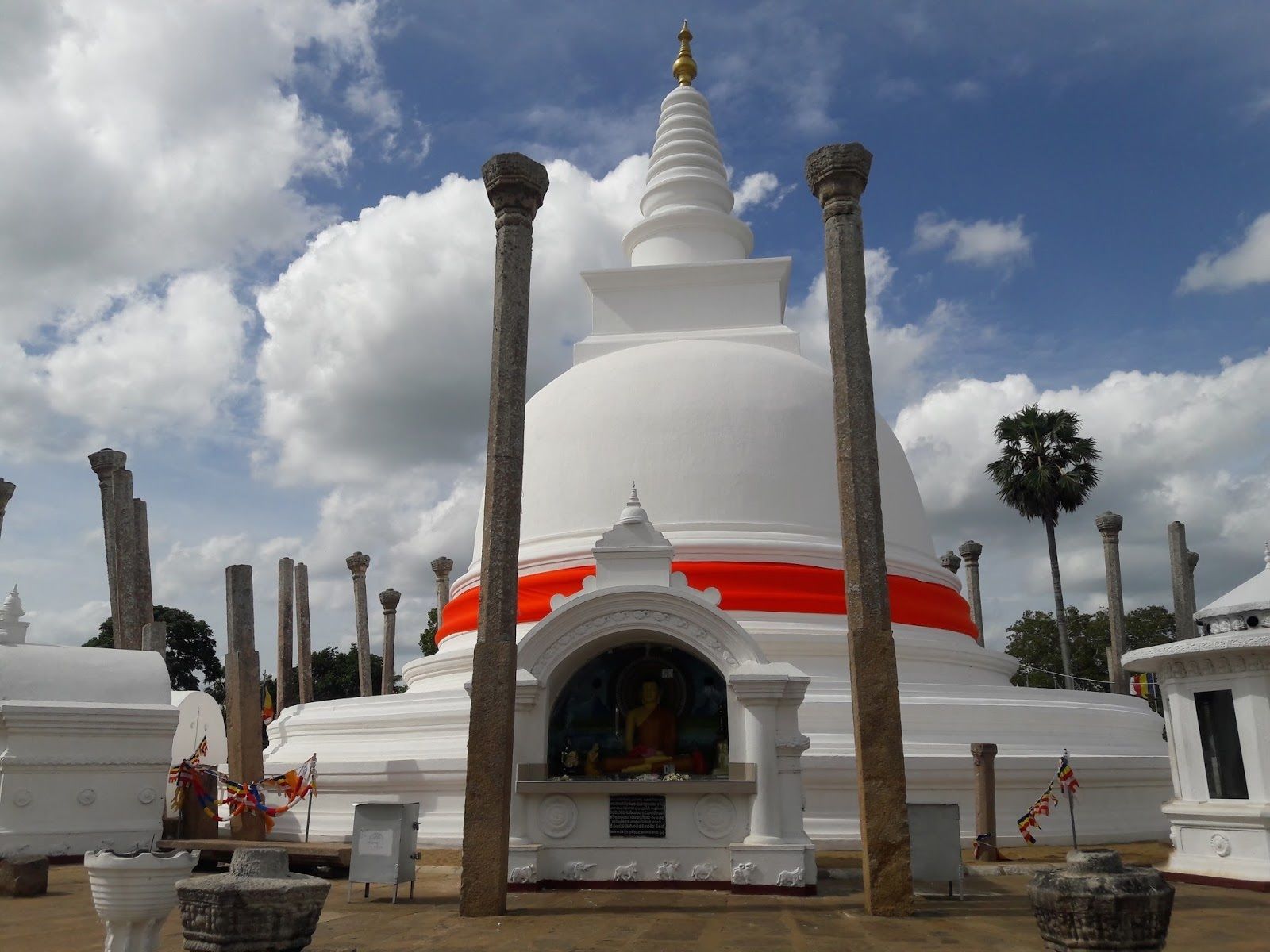 A white stupa with a red band, surrounded by stone pillars, set against a partly cloudy sky.