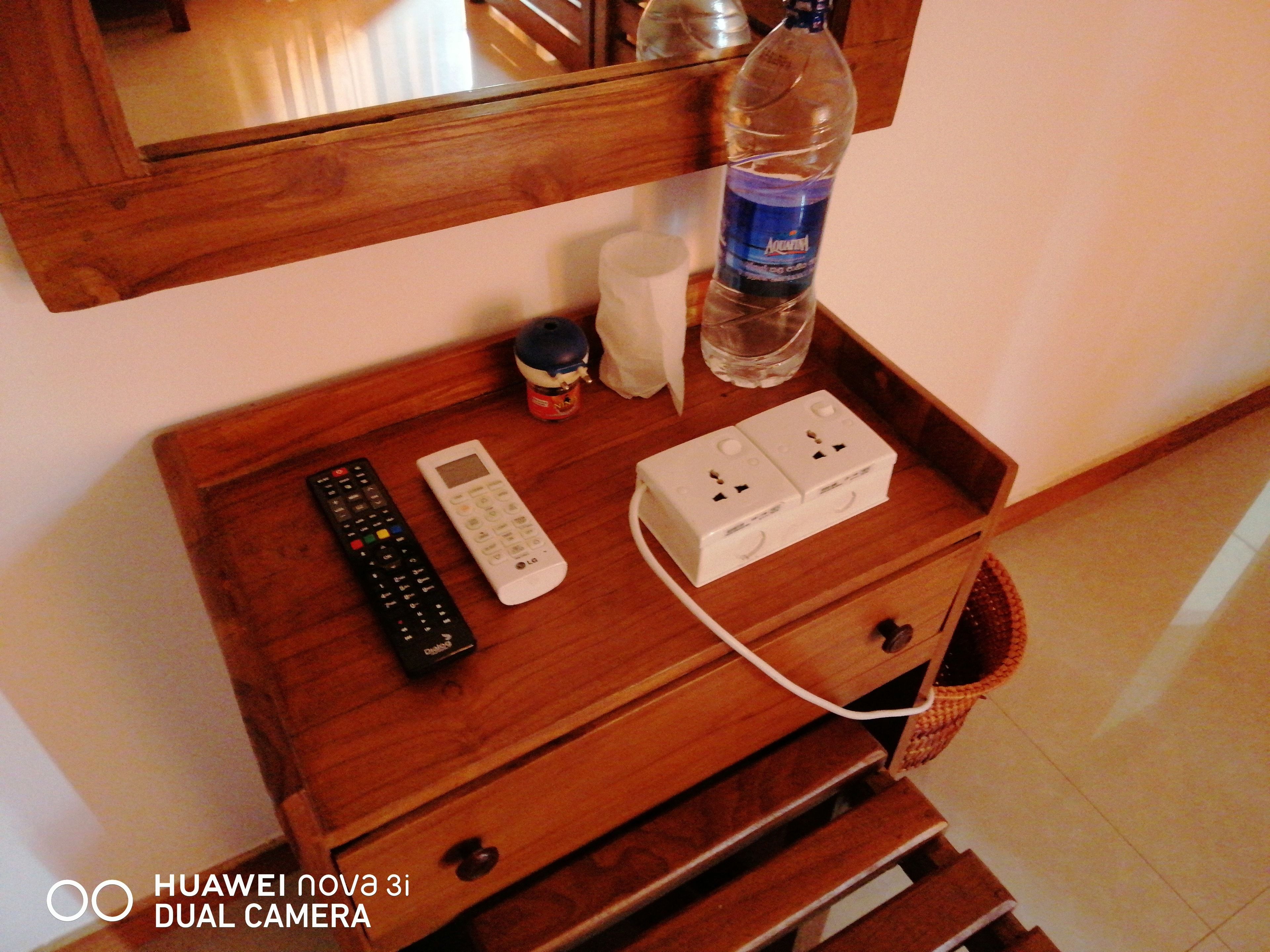 Un comodino in legno con un telecomando, una sveglia digitale, una ciabatta multipresa e una bottiglia d'acqua.