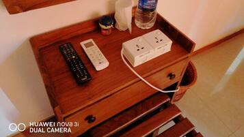 Un comodino in legno con un telecomando, una sveglia digitale, una ciabatta multipresa e una bottiglia d'acqua.