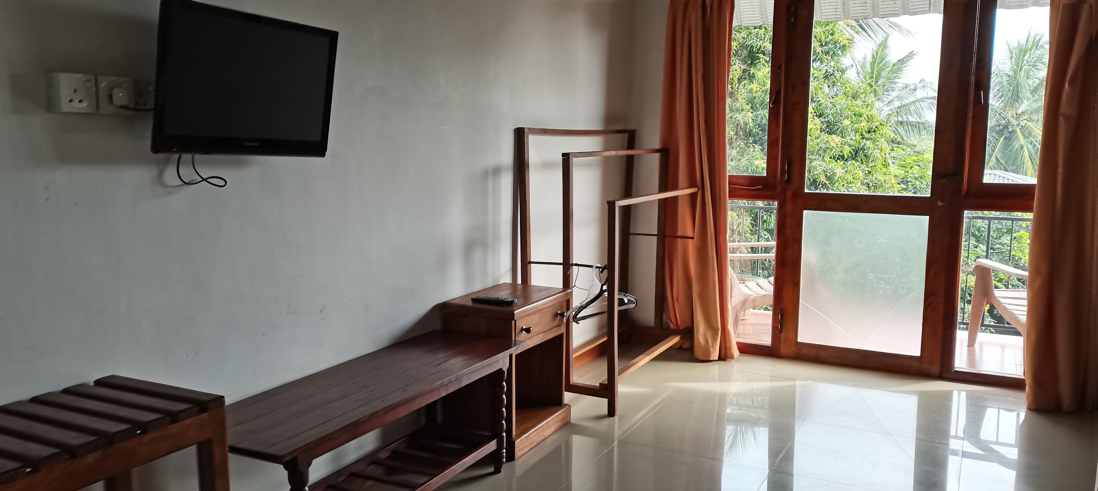 Una habitación con una televisión montada en la pared, un banco de madera, un perchero metálico y un balcón con cortinas.