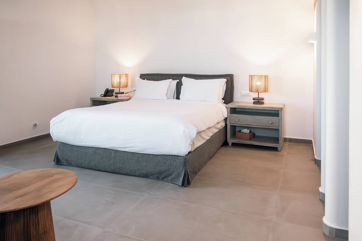 Una habitación de hotel moderna con una cama grande, dos mesitas de noche con lámparas, una pequeña mesa de madera y un banco gris.