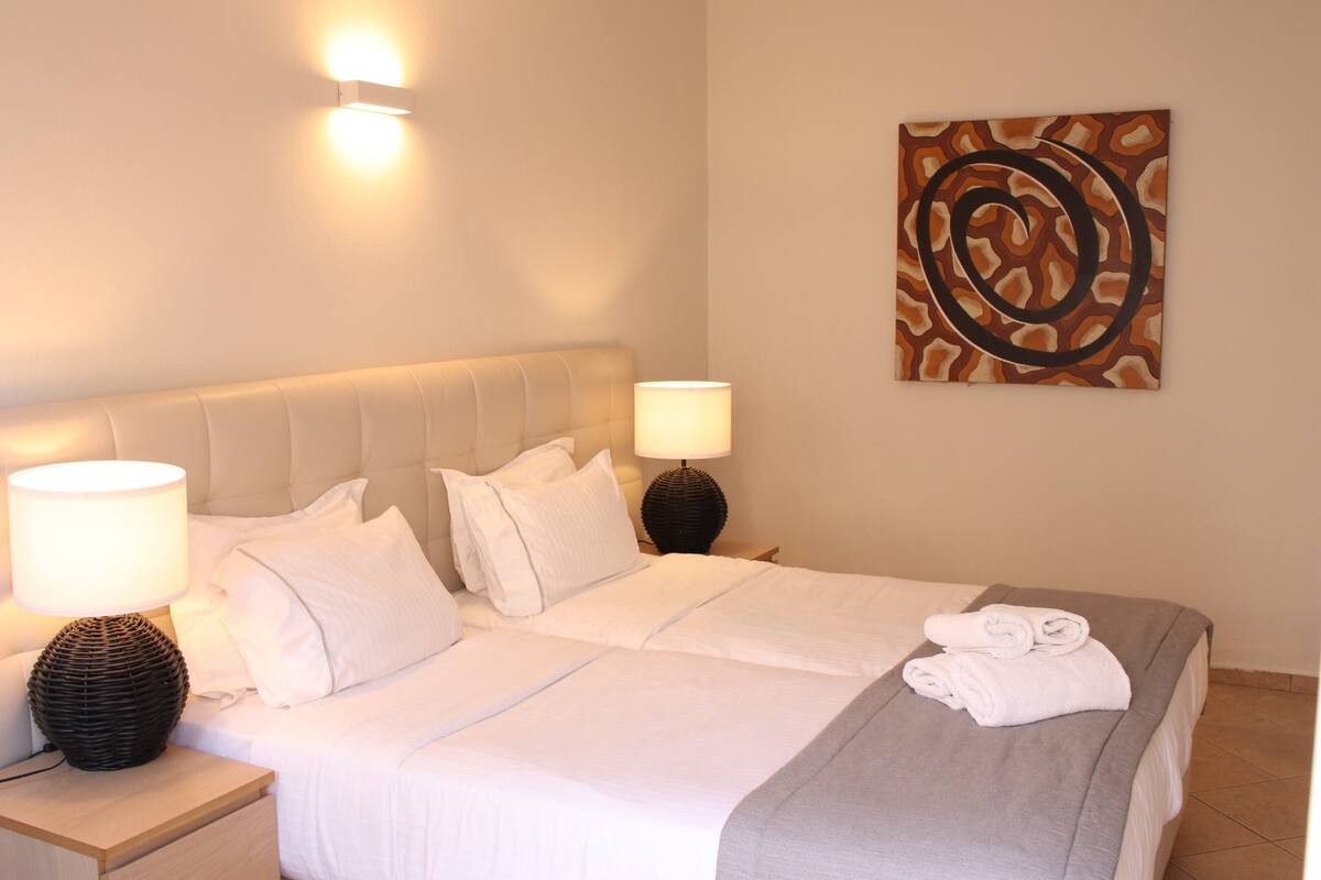 Una cama bien hecha con sábanas blancas, dos lámparas de noche y un cuadro en la pared.