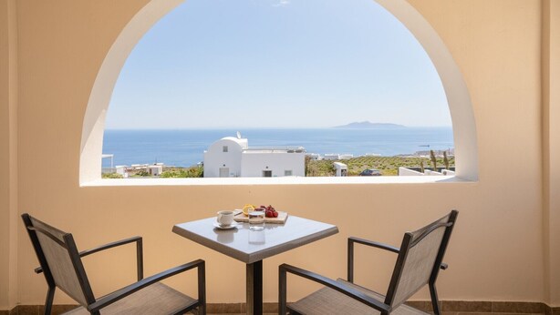 A balcony with a table and two chairs, overlooking a coastal view with buildings and the sea. - Villa Libertad (Santorini)