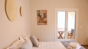 A bedroom with a bed, pillows, a painting, and a view of an outdoor area with a table and chairs. - Villa Libertad (Santorini)