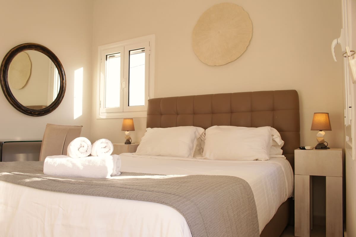 Habitación de hotel con una cama grande, dos lámparas de noche, un espejo redondo y una ventana con persianas.
