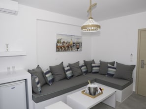 A modern living room with a sectional sofa, a glass coffee table, a painting on the wall, and a hanging light fixture. - Amaryllis Beach Hotel (Paros)