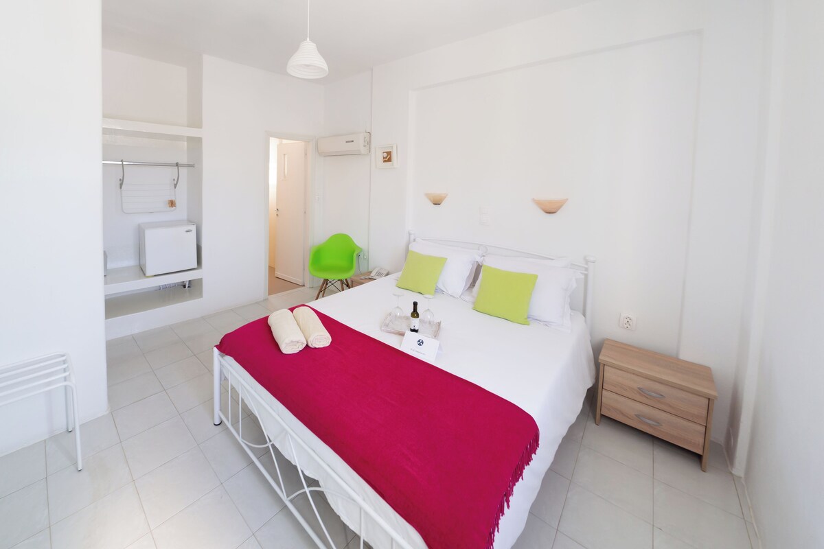 A modern hotel room with a bed, a small bedside table, a chair, a wardrobe, and a desk with a chair.