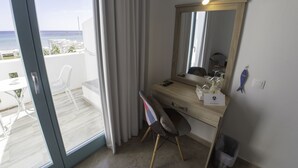 A hotel room with a large sliding glass door leading to a balcony with a view of the sea. There is a wooden desk with a mirror, a chair, and a small trash bin. - Amaryllis Beach Hotel (Paros)