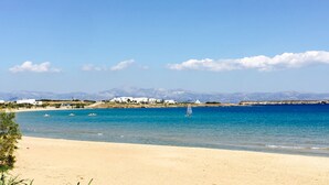 A beach with clear blue water, sandy shore, and white buildings in the background. - Amaryllis Beach Hotel (Paros)