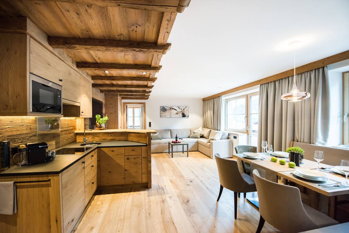 A modern kitchen with wooden beams, a dining area with a table and chairs, and a living space with a sofa and a TV.