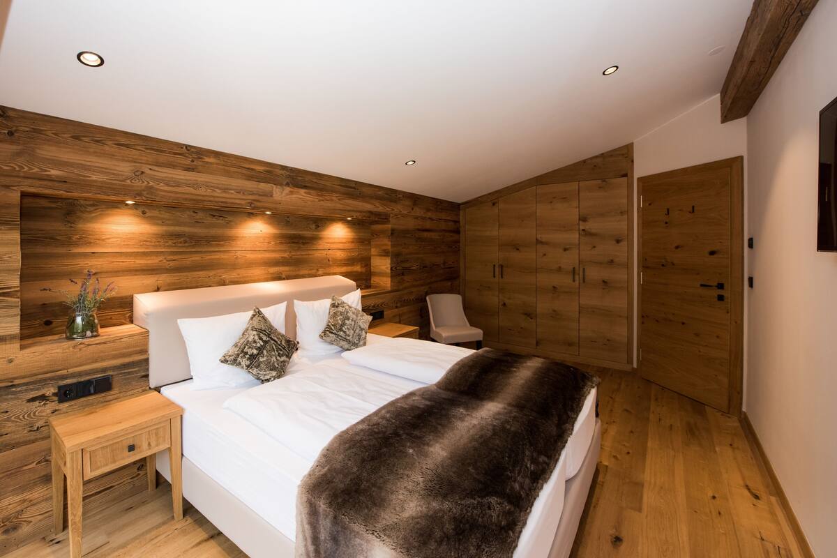 A bedroom with a wooden wall panel, a white bed, a nightstand, a chair, and a wardrobe.