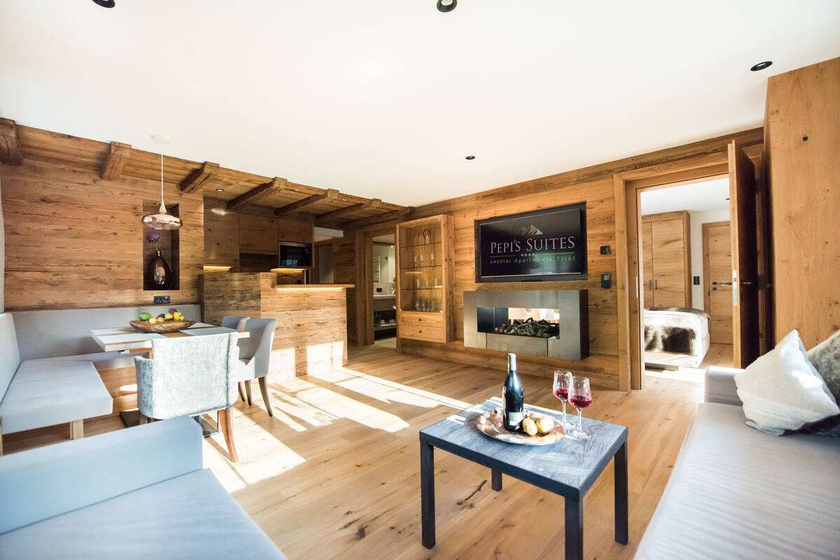 A cozy living room with a wooden interior, a sofa, a dining table with fruits and wine glasses, and a sign for 'Pepsi's Suites'.