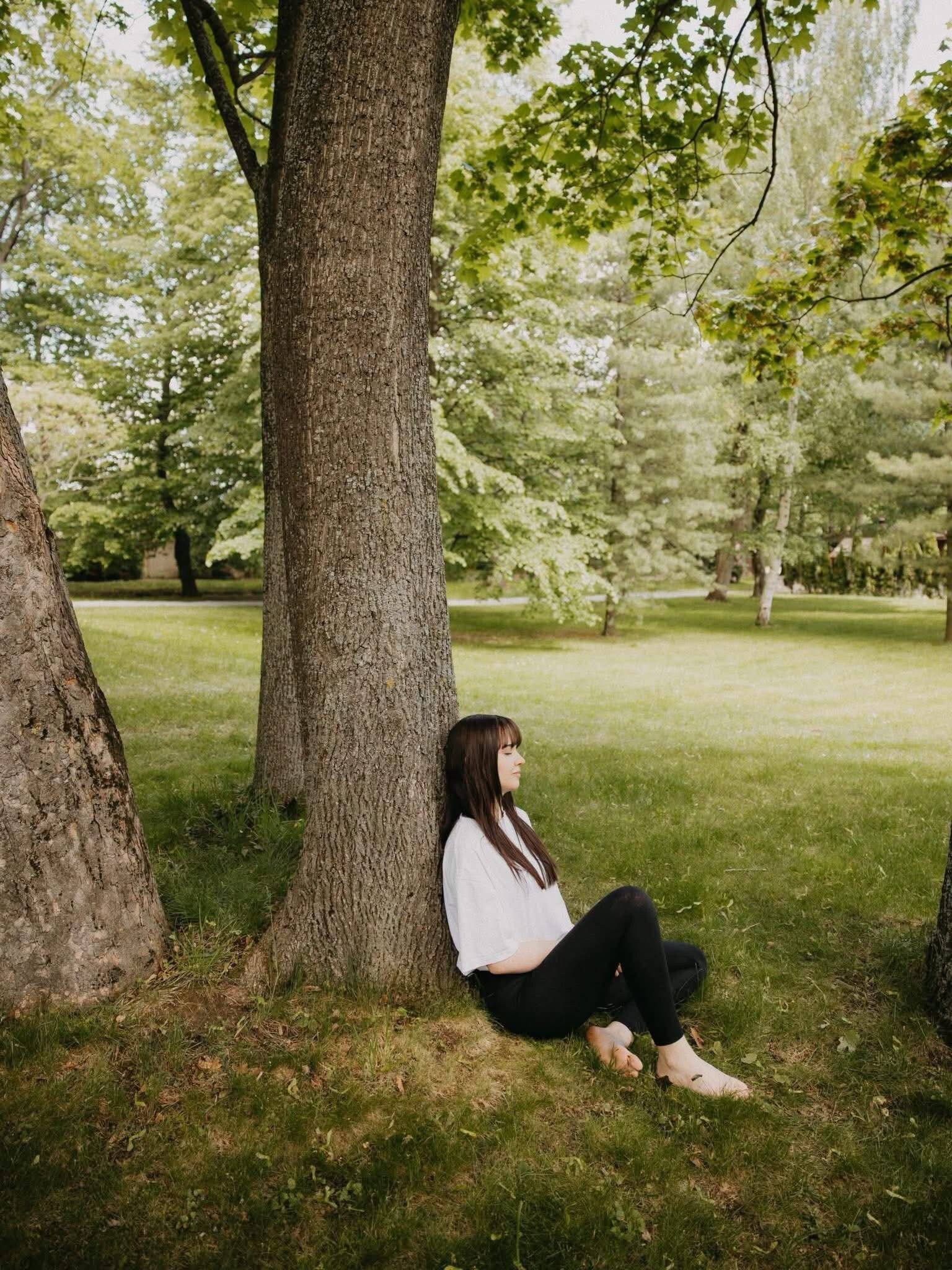 Eine Person sitzt auf der Wiese neben einem Baum in einem Park.