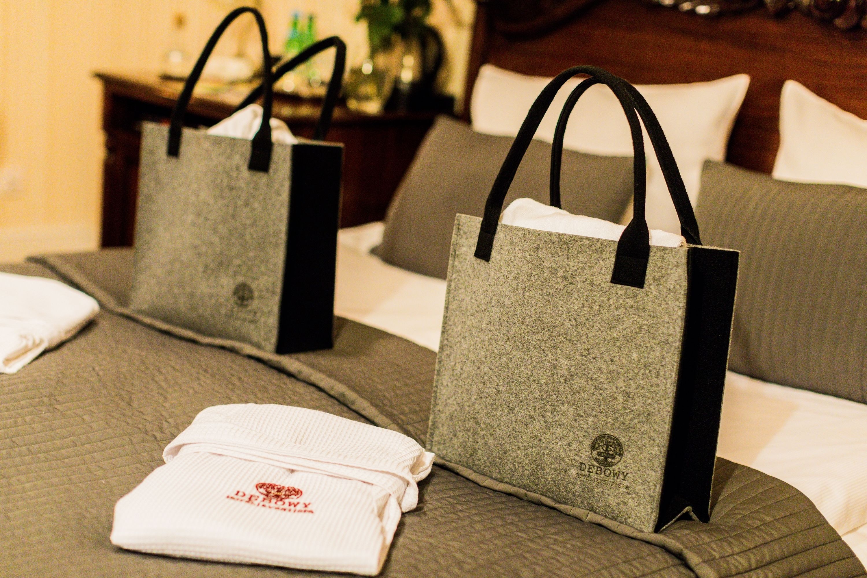 Two grey gift bags with black handles on a bed with a quilted cover and a neatly folded white towel with red text.
