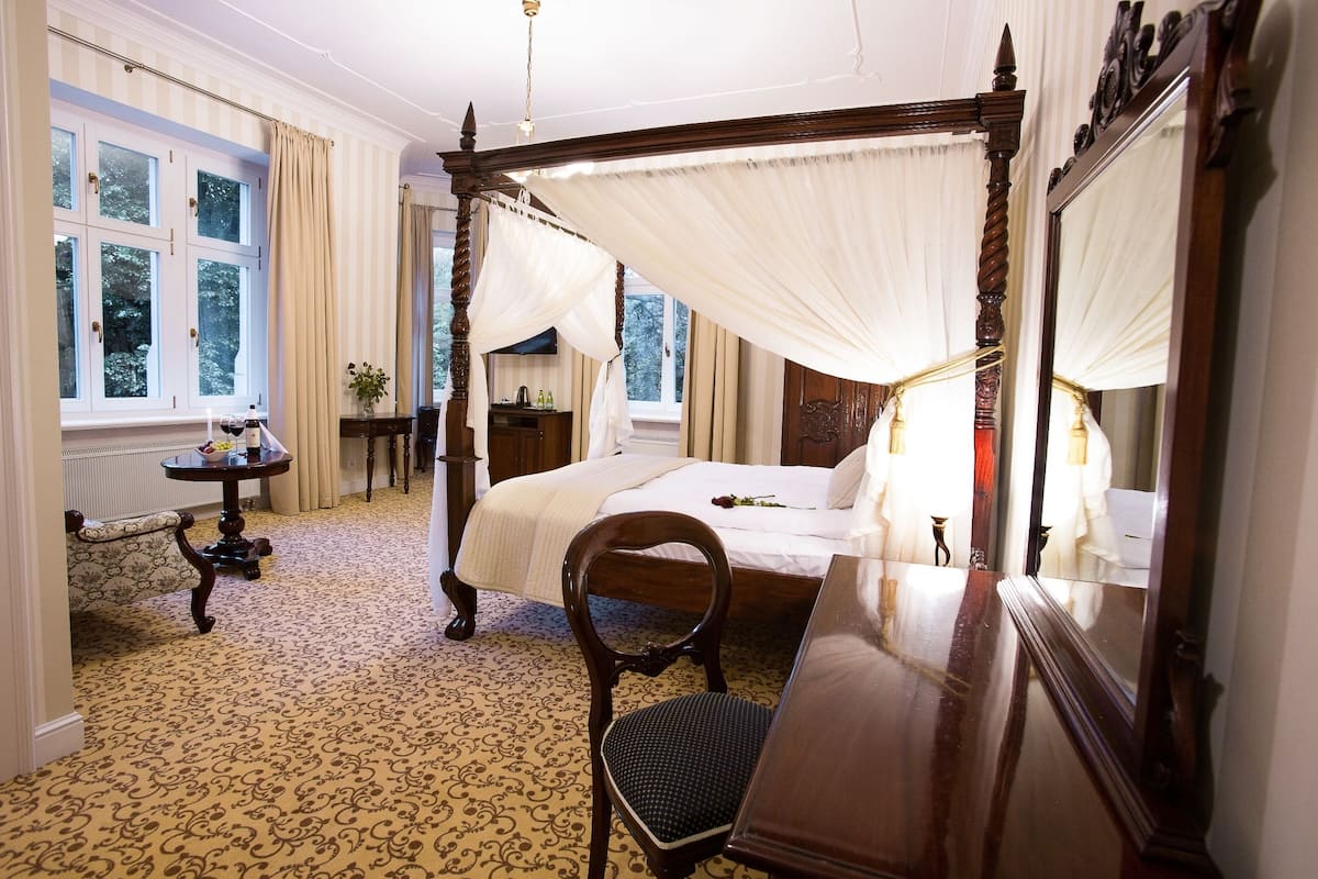 A four-poster canopy bed with a wooden frame, a round table with a vase of flowers, and a large mirror on the wall.