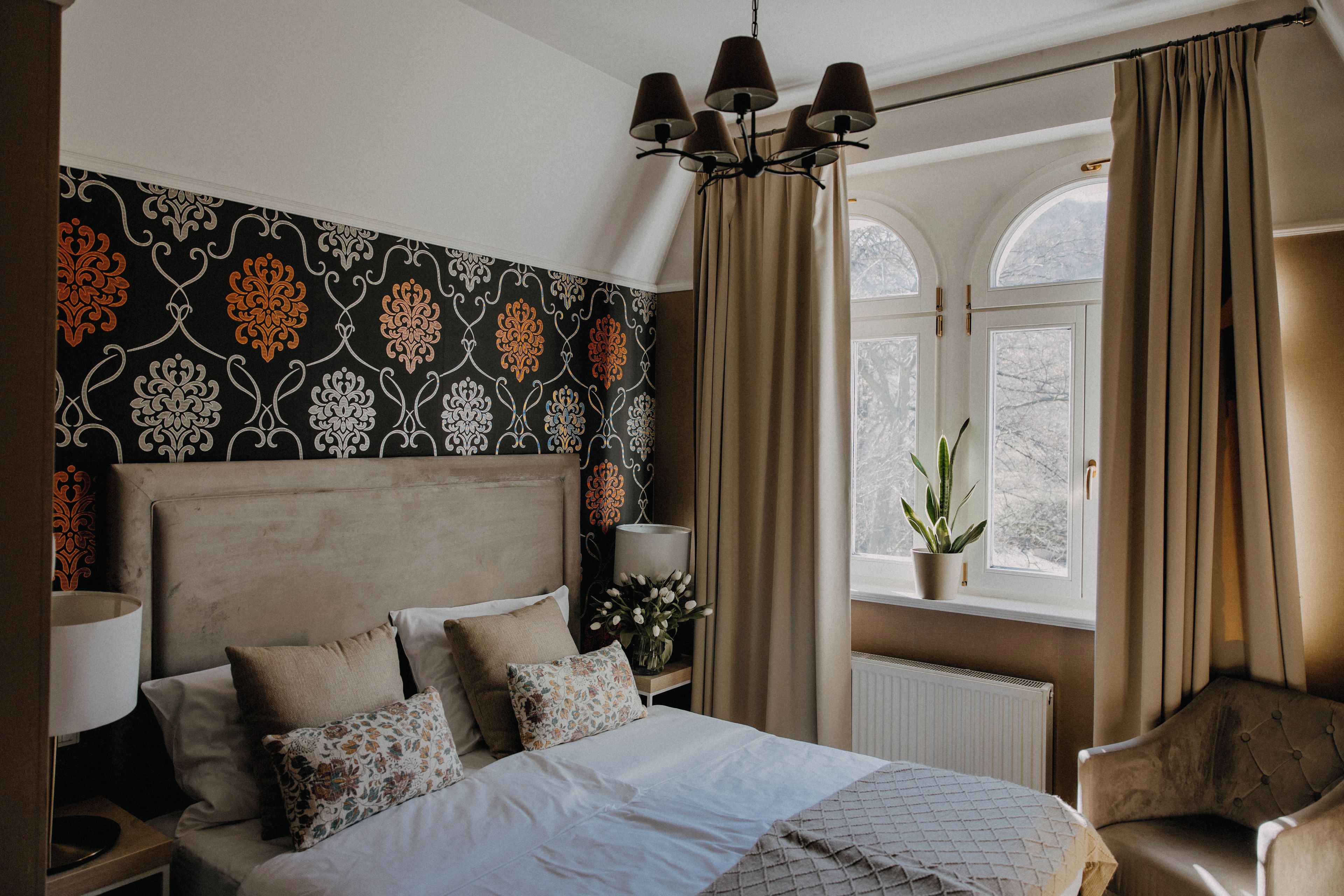 A bedroom with a bed, a nightstand, a lamp, a window with curtains, and a plant.