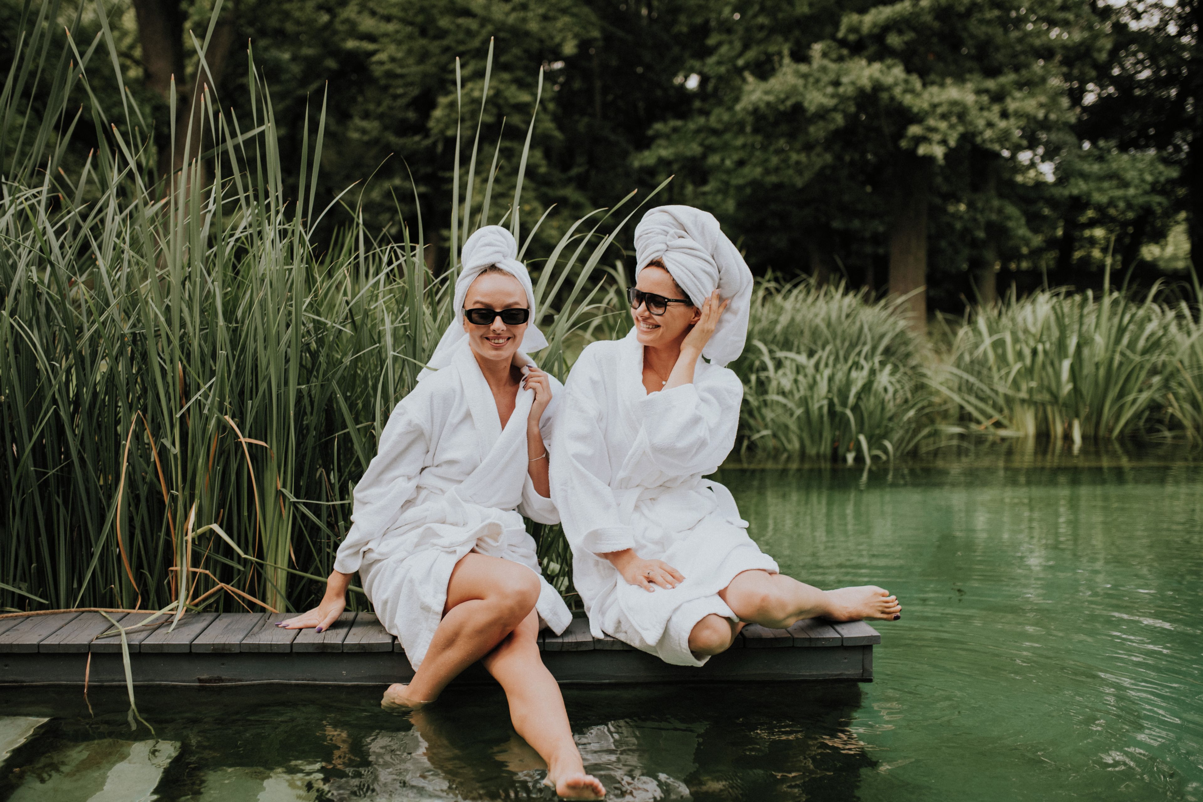 Two individuals seated on a wooden dock over water, surrounded by greenery.