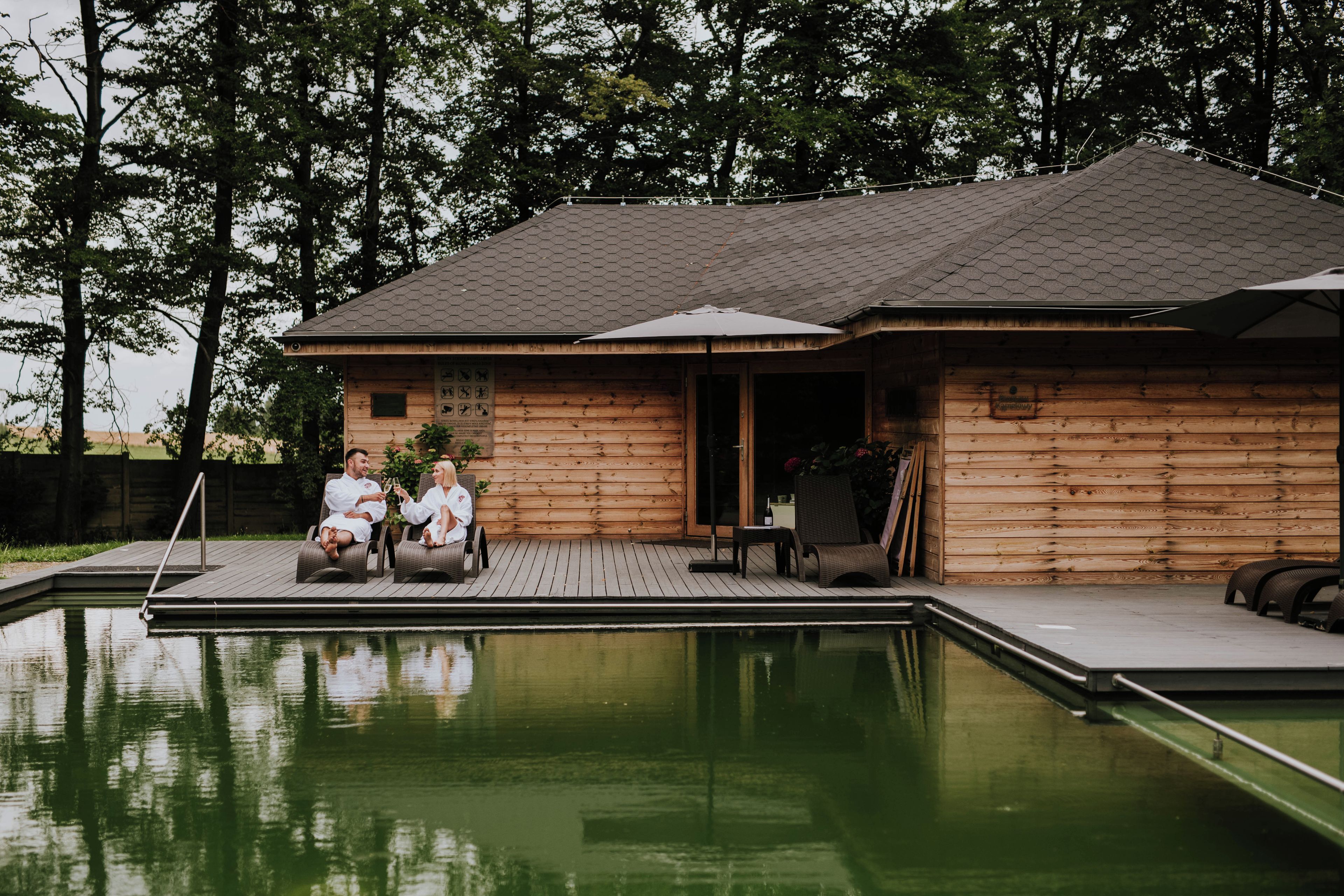 A wooden cabin with a pool, two people sitting on a deck chair, and a covered patio area.