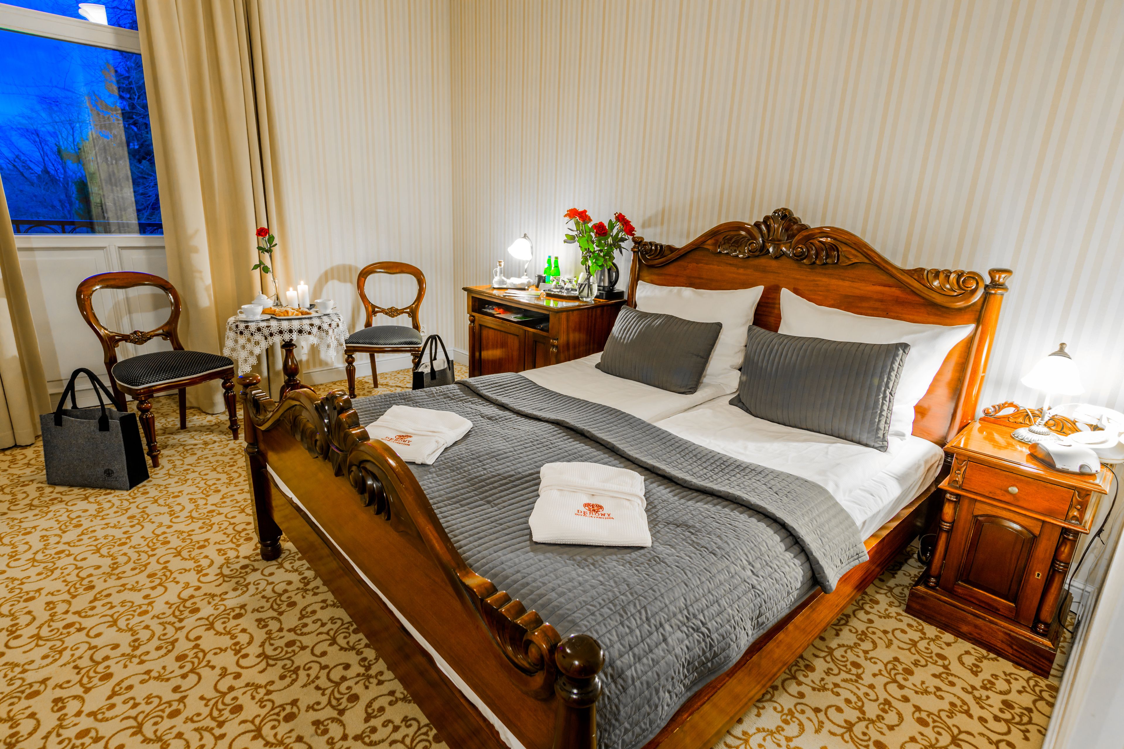 A hotel room with a large bed, a wooden headboard, a bedside table with a lamp, a small table with a vase of flowers, and a chair.