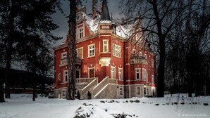 A large, red brick building with a steeply pitched roof and multiple chimneys, surrounded by snow-covered ground and trees.