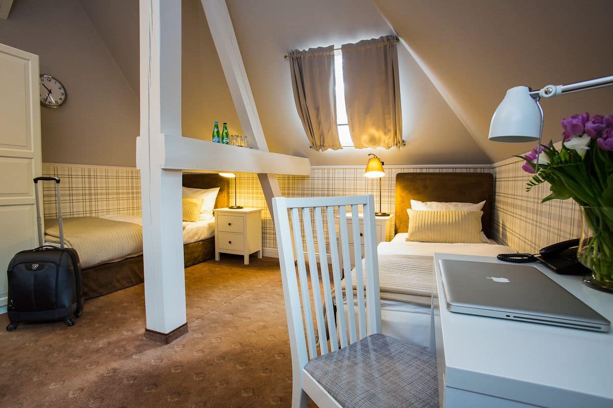 A hotel room with a bed, a desk with a laptop, a chair, a suitcase, and a nightstand with a lamp.