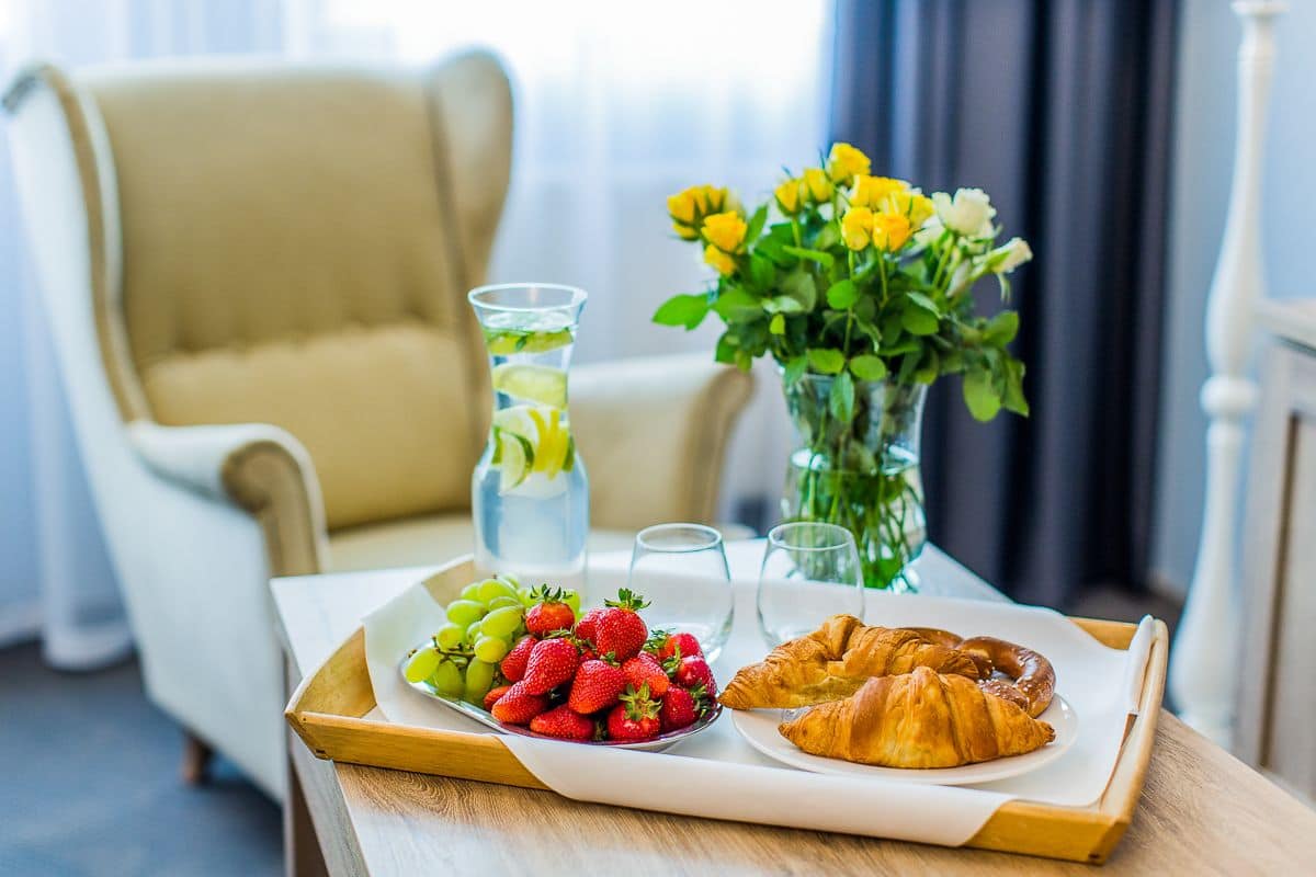 A breakfast tray with strawberries, grapes, and a croissant on a wooden table, a vase of yellow flowers, and a water bottle in the background.