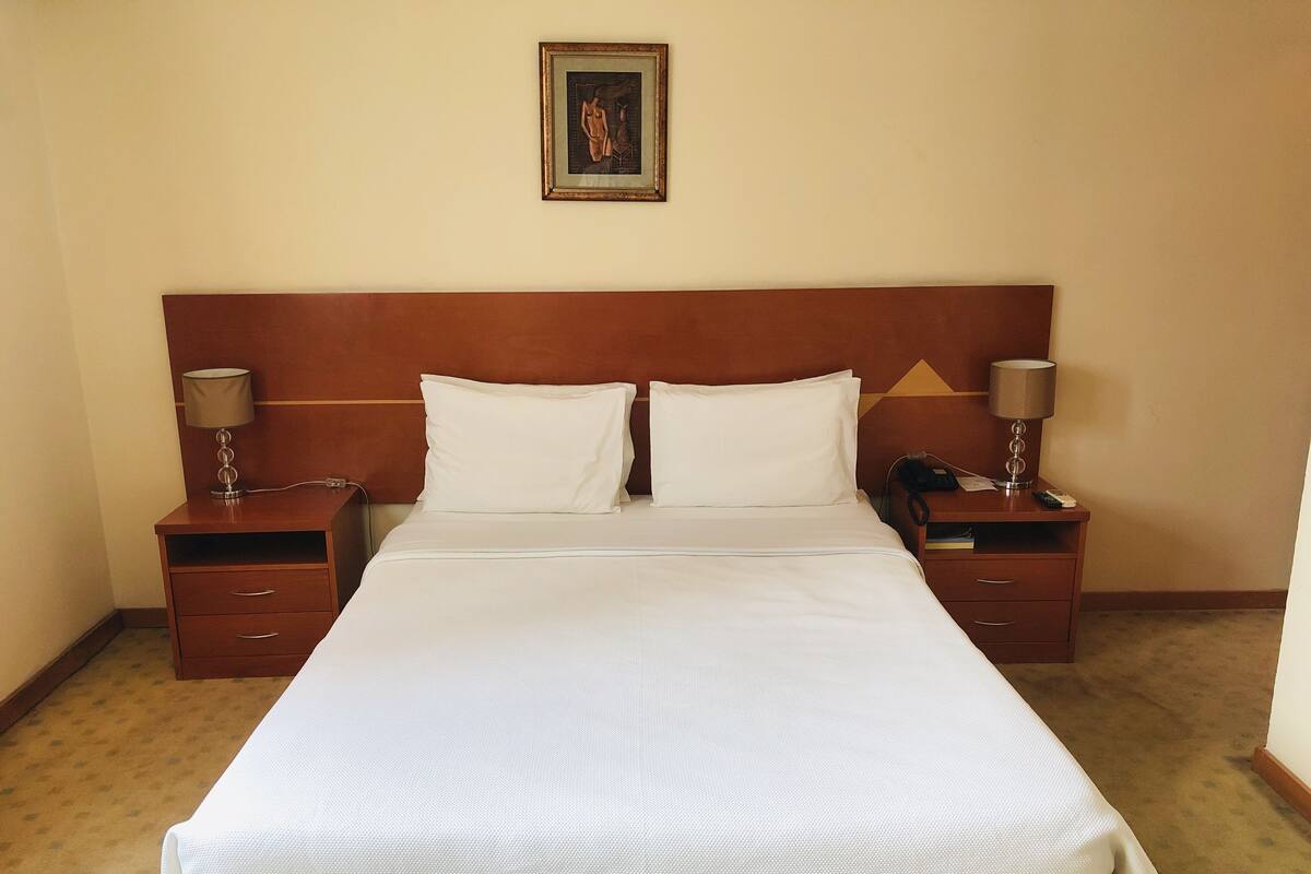Una cama bien hecha con sábanas blancas, dos mesitas de noche con lámparas y un cuadro en la pared.