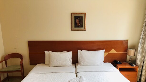 Une chambre d’hôtel avec un grand lit, deux oreillers, une tête de lit en bois, une table de chevet avec une lampe, une chaise et un tableau au mur.