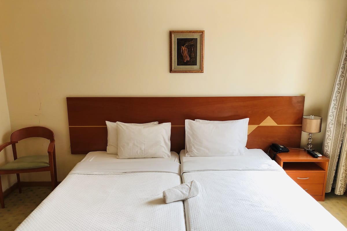 Habitación de hotel con una cama grande, dos almohadas, un cabecero de madera, una mesita de noche con una lámpara, una silla y un cuadro en la pared.