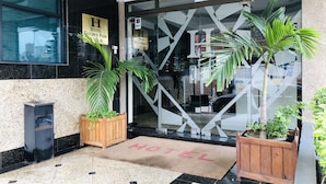 The entrance of the Hotel Golden Park with a modern glass facade, a wooden planter with a palm tree, and a doormat with "HOTEL" written on it. - Golden Park Hotel (Luanda)