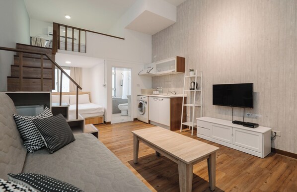 A compact living space with a sofa, a coffee table, a TV, a kitchen area, and a staircase leading to an upper level.
