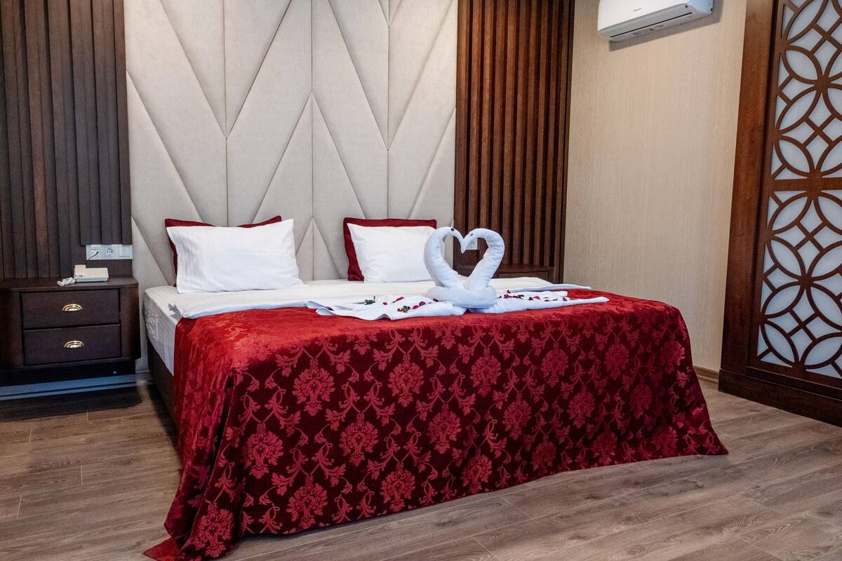 Cama bem arrumada com colcha vermelha e almofadas brancas, mesa de cabeceira de madeira com gaveta e um painel decorativo na parede.