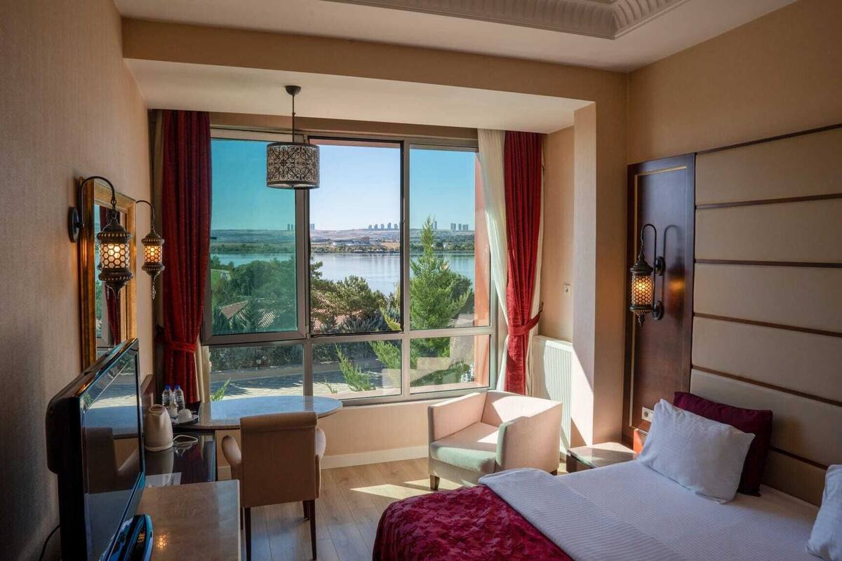 Quarto de hotel com uma janela grande, uma cama, uma escrivaninha e uma cadeira.