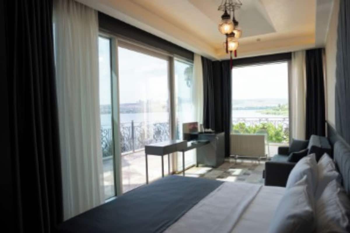 Quarto de hotel moderno com uma cama grande, uma escrivaninha e vista da cidade através de janelas amplas.
