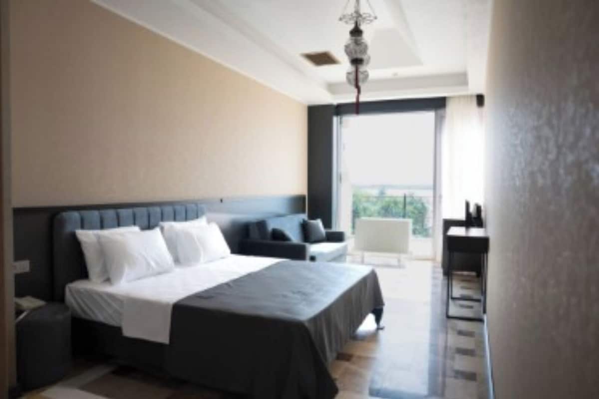 Quarto de hotel moderno com uma cama grande, área de estar com sofá e poltronas, uma mesinha e varanda com vista.